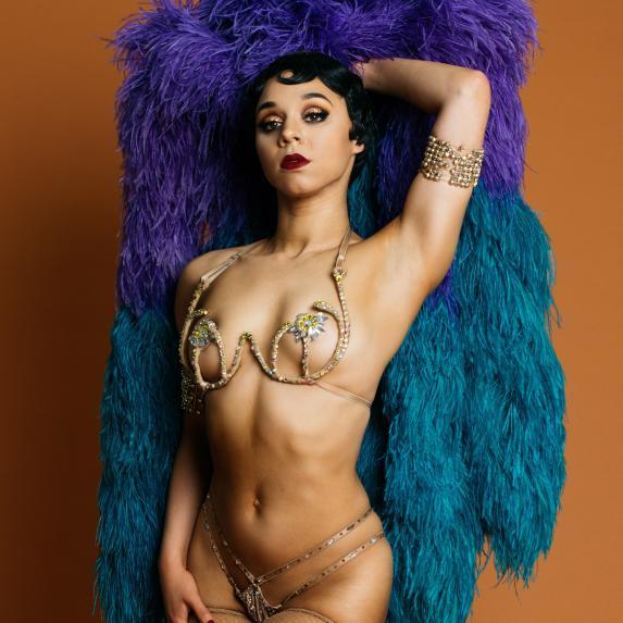 Demi Noire, posing in showgirl costume, a crystalled wire bra, pasties, a rhinestoned thong, and a matching arm cuff. Her black hair is short and finger-waved. She is holding up a purple and teal feather boa behind her.
