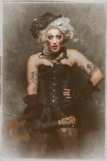 Lena Mae Lenman as her Music Hall, Victorian persona, 'Aunty Mae.' She is a very Dickensian looking character, with very bold makeup, messy blonde hair in an Edwardian-style updo, wearing a black corset, black French knickers, holey black tights, and black fingerless gloves. She us holding a ukulele and staring us down through the lens.
